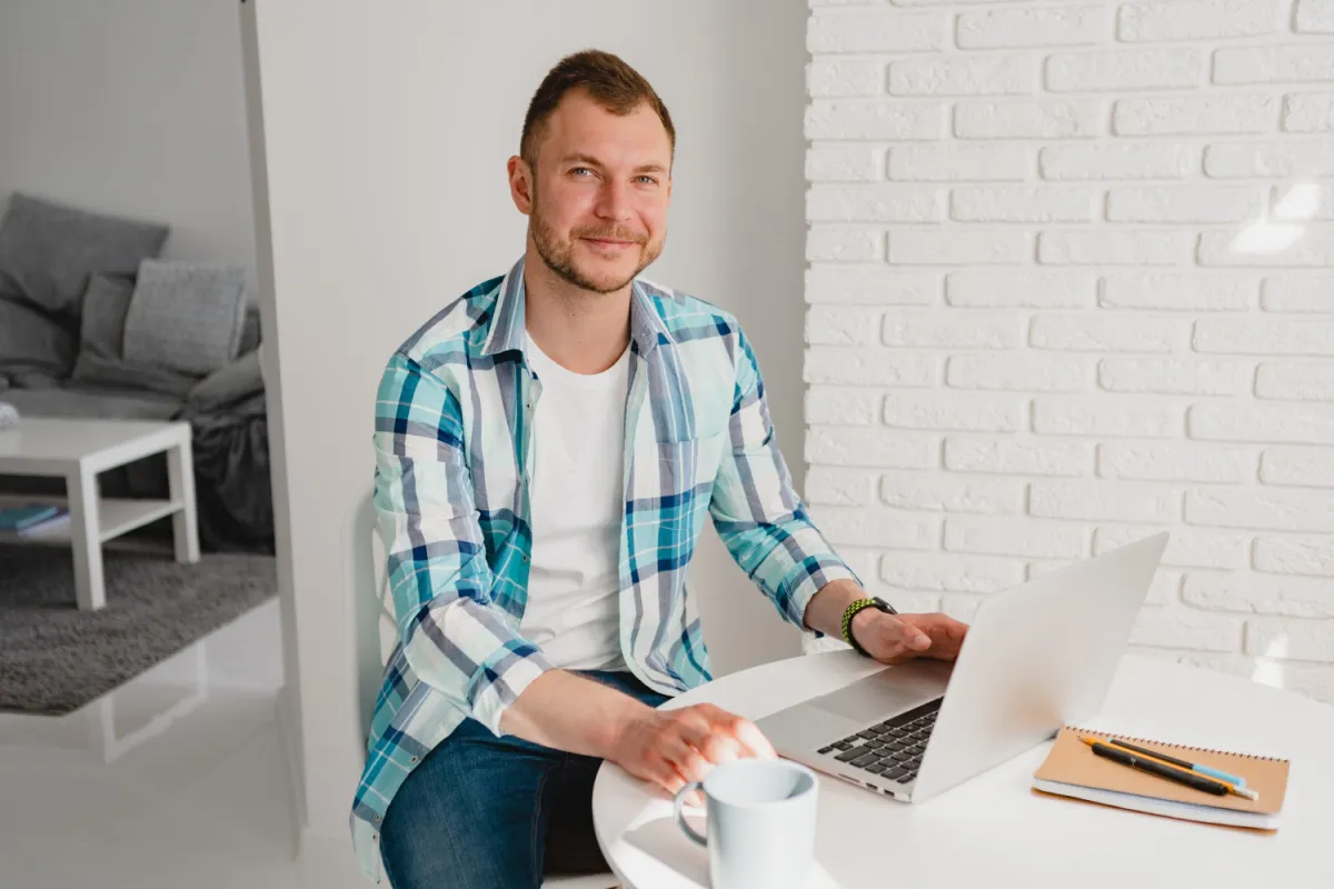 Handsome smiling man in shirt sitting in kitchen at home at table working online on laptop from hom