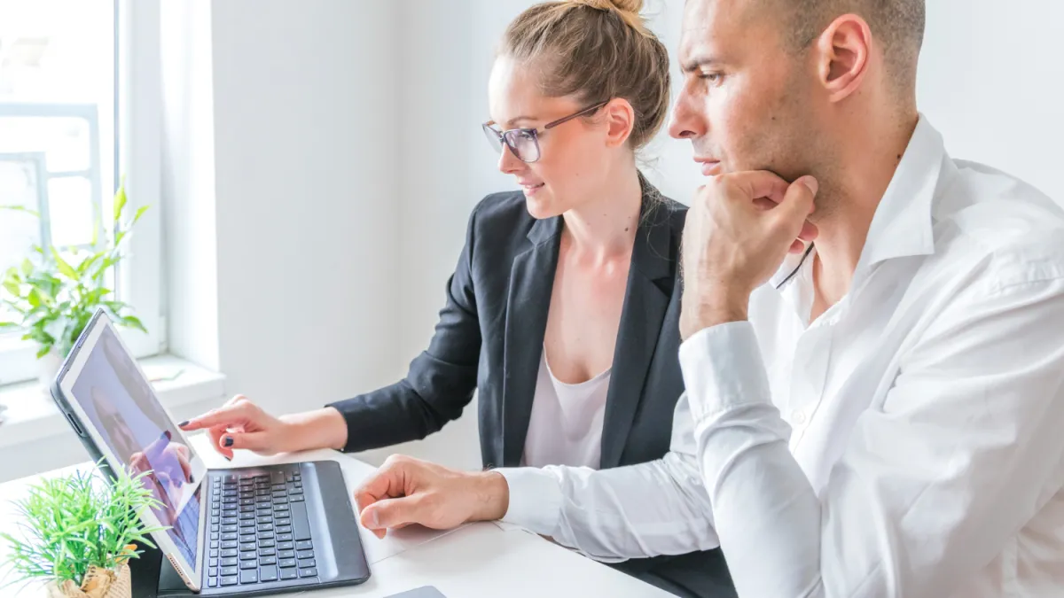 irodai környezetben és öltözetben laptopot néz egy férfi és egy nő