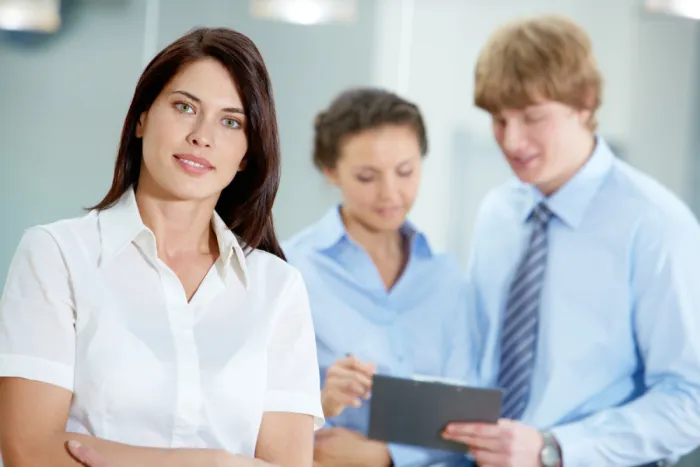 Fiatal dolgozó nő a kamerába néz érdeklődően, munkatársai a háttérben