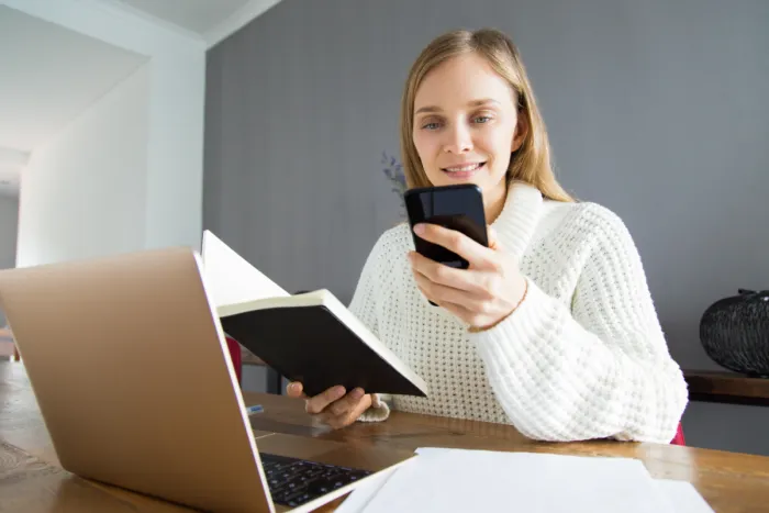 mosolygó hölgy laptop előtt mobiltelefonnal és jegyzetfüzettel